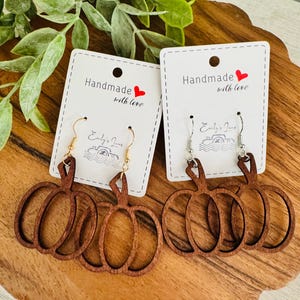 May include: Two pairs of wooden pumpkin earrings on white cards that read "Handmade with love." The earrings are brown with gold and silver hooks. The earrings are displayed on a wooden surface with green foliage.