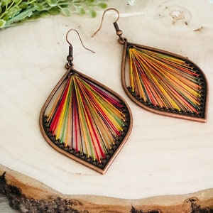 May include: A pair of teardrop-shaped earrings with a copper-toned metal frame and colourful thread woven into the design. The earrings have a bohemian style.