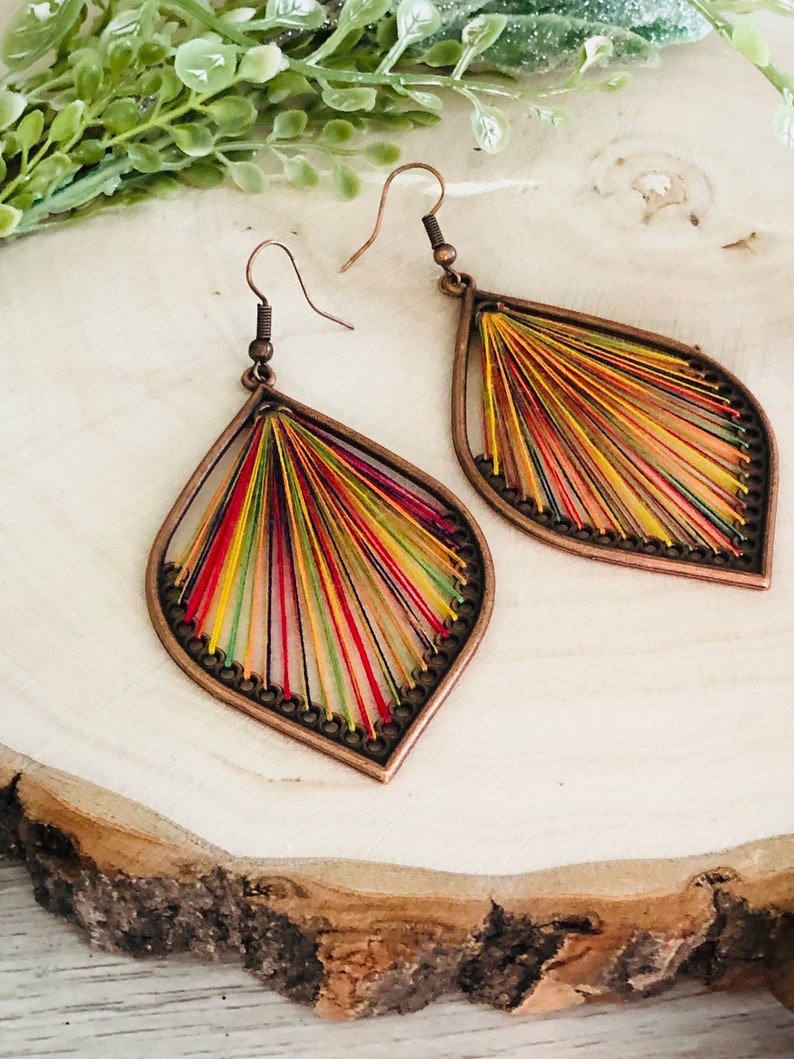 May include: A pair of teardrop-shaped earrings with a copper-toned metal frame and colourful thread woven into the design. The earrings have a bohemian style.