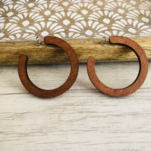 Puede incluir: Un par de pendientes de aro hechos de madera de color marr&oacute;n oscuro. Cada pendiente presenta un dise&ntilde;o circular abierto con un acabado liso y un cierre de poste de metal plateado para una fijaci&oacute;n segura al l&oacute;bulo.
