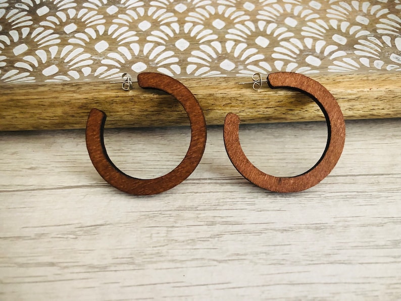Puede incluir: Un par de pendientes de aro hechos de madera de color marr&oacute;n oscuro. Cada pendiente presenta un dise&ntilde;o circular abierto con un acabado liso y un cierre de poste de metal plateado para una fijaci&oacute;n segura al l&oacute;bulo.