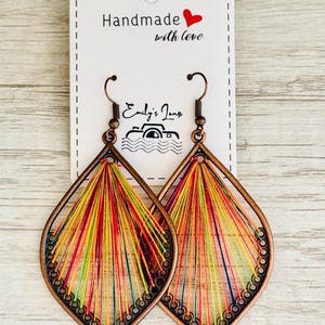 May include: A pair of handmade teardrop-shaped earrings. The earrings are made from copper wire and decorated with colourful threads. The earrings are hanging from a white card with the text "Handmade with love" and a red heart.