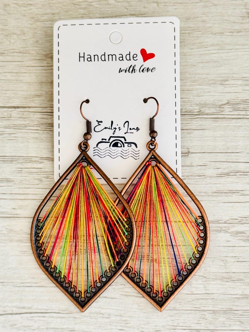 May include: A pair of handmade teardrop-shaped earrings. The earrings are made from copper wire and decorated with colourful threads. The earrings are hanging from a white card with the text "Handmade with love" and a red heart.