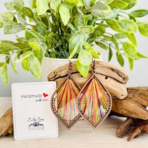 May include: A pair of colourful, handmade earrings with a copper wire frame and a rainbow of threads woven into a teardrop shape. The earrings are on a wooden surface with a small white card that says "Handmade with love" and "Emily Jane's".