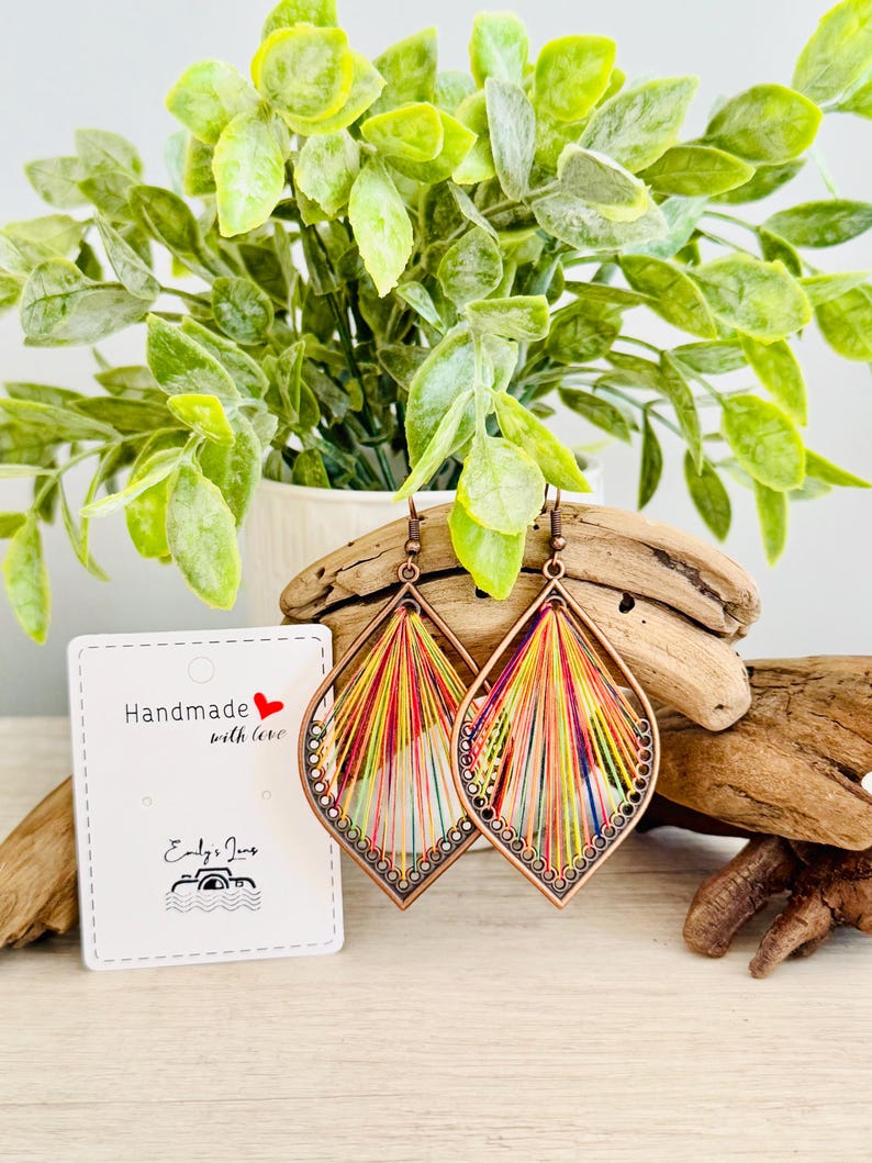 May include: A pair of colourful, handmade earrings with a copper wire frame and a rainbow of threads woven into a teardrop shape. The earrings are on a wooden surface with a small white card that says "Handmade with love" and "Emily Jane's".