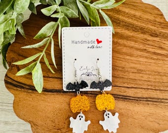 May include: Dangle earrings featuring a black glitter bat, an orange glitter pumpkin, and a white ghost charm. The earrings are attached to a white display card with the text Handmade with love and Emily's Jewelry printed in black ink.