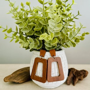 May include: A pair of wooden earrings with a rectangular geometric design. The earrings are a warm brown color and hang from small rectangular wooden posts. The earrings are displayed in front of a white vase with green foliage.