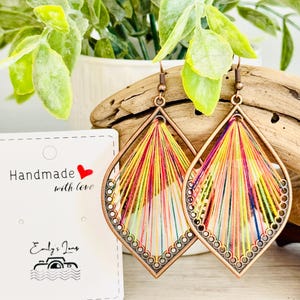 May include: A pair of colourful, handmade earrings with a copper-toned metal frame. The earrings are made with a variety of colourful threads woven together in a geometric pattern. The earrings are hanging from a wooden surface.