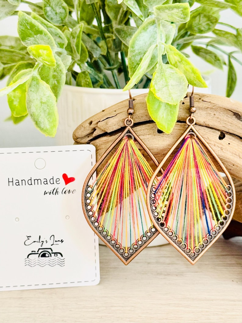 May include: A pair of colourful, handmade earrings with a copper-toned metal frame. The earrings are made with a variety of colourful threads woven together in a geometric pattern. The earrings are hanging from a wooden surface.