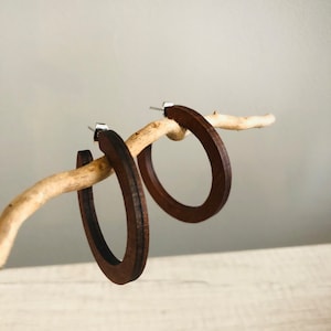 Puede incluir: Un par de pendientes de aro de madera de color marr&oacute;n oscuro con un acabado liso y plano y postes de metal plateado. Los aros circulares presentan una textura de veta de madera visible y cuelgan de una ramita clara.