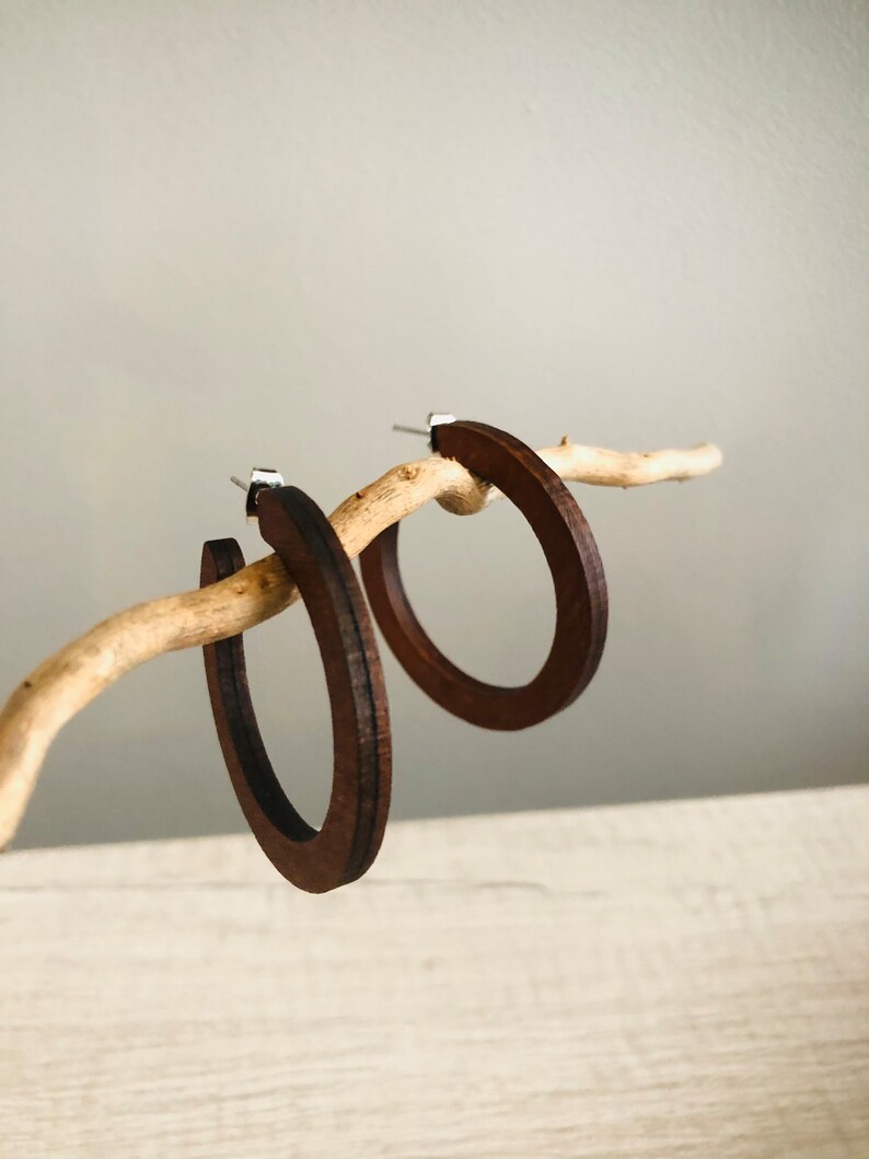 Puede incluir: Un par de pendientes de aro de madera de color marr&oacute;n oscuro con un acabado liso y plano y postes de metal plateado. Los aros circulares presentan una textura de veta de madera visible y cuelgan de una ramita clara.