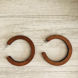 Puede incluir: Un par de pendientes de aro circulares hechos de madera de color marr&oacute;n oscuro con una textura de grano visible. Cada pendiente cuenta con un poste de metal plateado y un cierre de mariposa, formando un dise&ntilde;o de aro abierto.