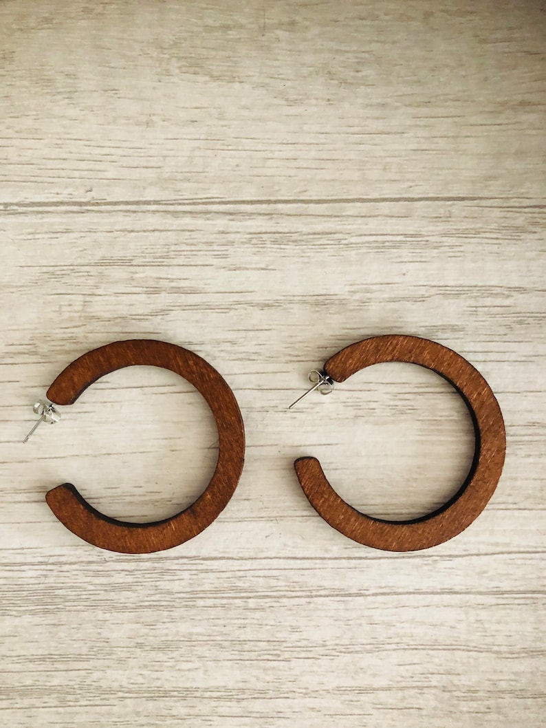 Puede incluir: Un par de pendientes de aro circulares hechos de madera de color marr&oacute;n oscuro con una textura de grano visible. Cada pendiente cuenta con un poste de metal plateado y un cierre de mariposa, formando un dise&ntilde;o de aro abierto.
