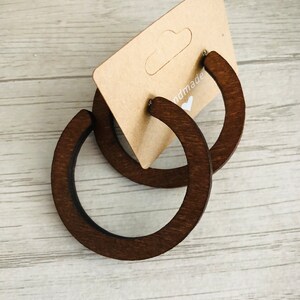 Puede incluir: Un par de aros de madera de color marr&oacute;n oscuro sujetos a una tarjeta de presentaci&oacute;n de cart&oacute;n color canela. La tarjeta muestra el texto parcial "handmade" seguido de un peque&ntilde;o icono de coraz&oacute;n blanco. Los aros son circulares y lisos.