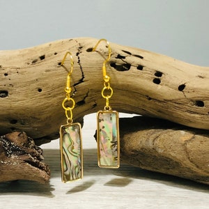 May include: Gold dangle earrings with a rectangular abalone shell inlay. The shell has a pearlescent iridescent finish with shades of pink, green, and blue.