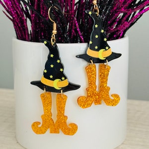 May include: A pair of witch-themed earrings with black witch hats and gold glitter boots. The hats have yellow stars and a crescent moon.