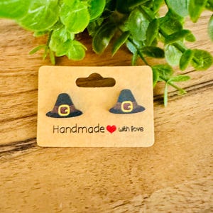 May include: A pair of witch hat earrings on a brown card. The earrings are black with a gold buckle. The card reads "Handmade with love" in black text with a red heart. The background is a wooden surface with green leaves.