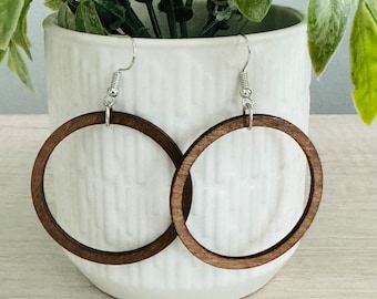 Puede incluir: Un par de pendientes de aro circulares hechos de madera de color marrón oscuro. Cada pendiente presenta un anillo de madera fino y plano suspendido de un gancho de metal plateado. El diseño es sencillo y muestra la veta de la madera.