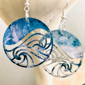 May include: A pair of silver earrings with a blue and white wave design. The earrings are made of acrylic and have a circular shape.