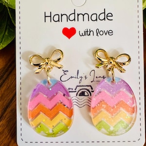 May include: A pair of handmade Easter egg earrings. Each earring features a gold bow at the top and a colorful, chevron-patterned egg pendant with glitter accents. The card reads "Handmade with love."