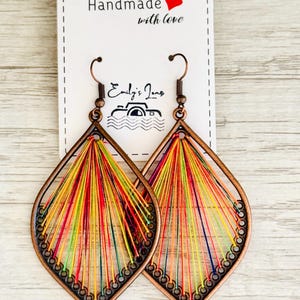 May include: A pair of handmade earrings in a teardrop shape. The earrings are made from copper wire and feature a colourful thread design. The earrings are labelled "Handmade with love".