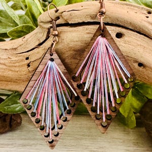 May include: A pair of diamond-shaped wooden earrings featuring a series of small circular cutouts along the bottom edges. Each earring is threaded with thin, radiating strands of pink, white, and light blue embroidery floss in a fan pattern.
