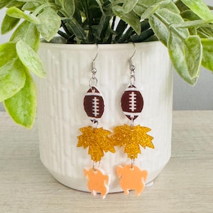 May include: A pair of Thanksgiving-themed earrings. Each earring features a brown football, a gold glitter leaf, and an orange turkey. The earrings are suspended from silver-colored fish hook ear wires. The earrings are displayed in front of a white pot with green foliage.