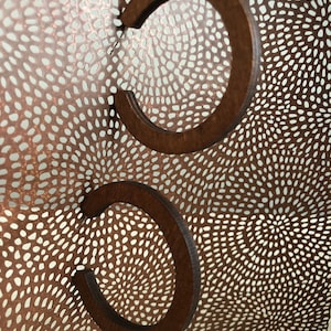 Puede incluir: Dos pendientes de aro de madera marr&oacute;n oscuro con un dise&ntilde;o circular abierto. Los pendientes est&aacute;n colocados frente a una l&aacute;mina met&aacute;lica curva de color cobre con un patr&oacute;n denso y complejo de peque&ntilde;os recortes alargados.