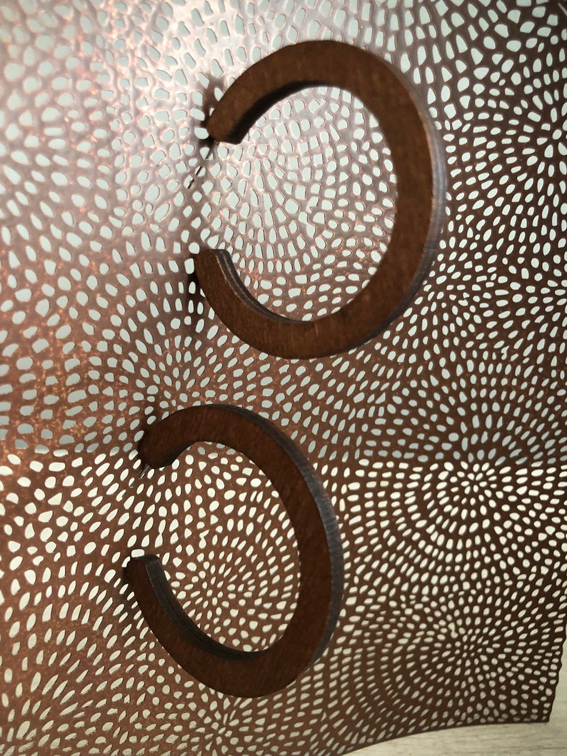 Puede incluir: Dos pendientes de aro de madera marr&oacute;n oscuro con un dise&ntilde;o circular abierto. Los pendientes est&aacute;n colocados frente a una l&aacute;mina met&aacute;lica curva de color cobre con un patr&oacute;n denso y complejo de peque&ntilde;os recortes alargados.