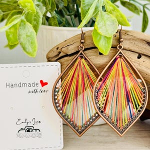 May include: A pair of colourful, handmade earrings with a teardrop shape. The earrings are made of copper wire and colourful threads. The earrings are hanging from a wooden surface.