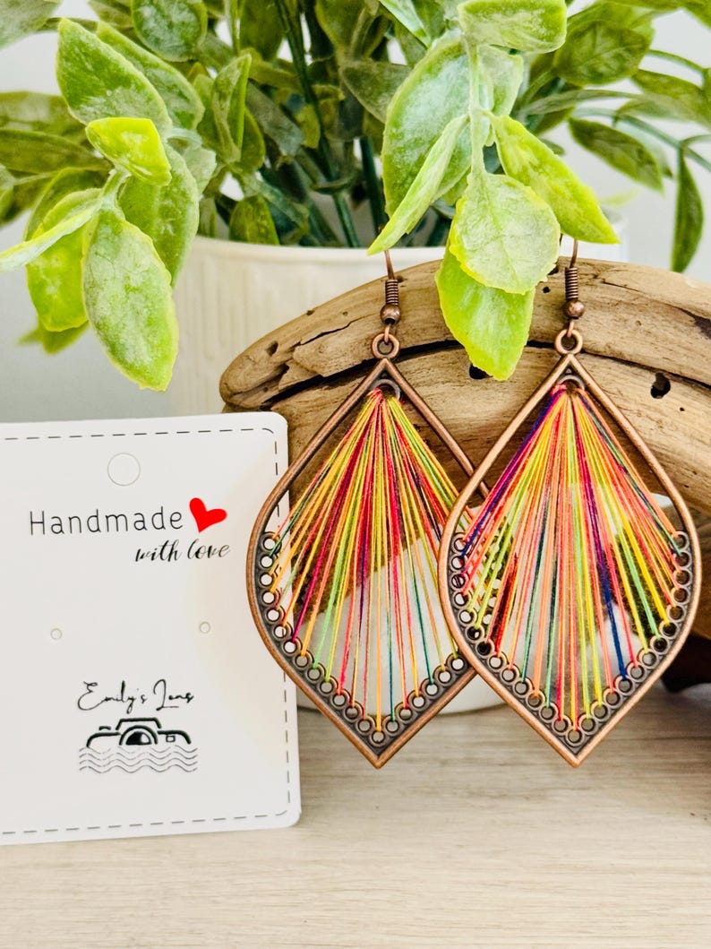May include: A pair of colourful, handmade earrings with a teardrop shape. The earrings are made of copper wire and colourful threads. The earrings are hanging from a wooden surface.