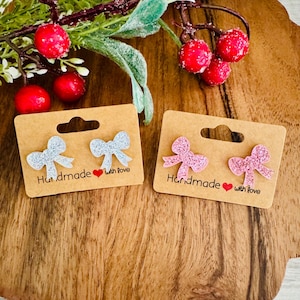May include: Two pairs of bow-shaped earrings on brown cardboard tags. One pair is silver glitter, the other is pink glitter. Each tag reads "Handmade with love" in black text with a red heart.