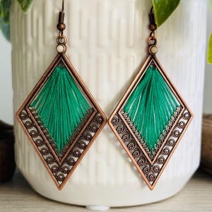 May include: A pair of green and copper diamond-shaped earrings with a delicate, intricate design. The earrings feature a green thread tassel hanging from the center of each diamond.