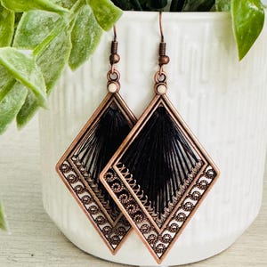 May include: A pair of copper-colored diamond-shaped earrings with black thread detailing. The earrings have a decorative border with a floral pattern and are suspended from copper-colored hooks.