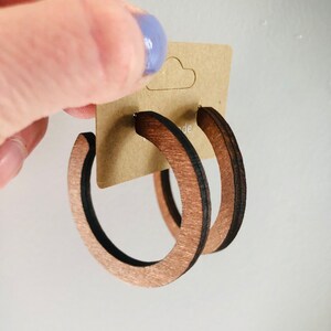 Puede incluir: Un par de pendientes de aro circulares hechos de madera de color marr&oacute;n oscuro con una textura de grano visible. Los pendientes est&aacute;n sujetos a una tarjeta de presentaci&oacute;n de cart&oacute;n color canela con una palabra parcial que termina en ade.
