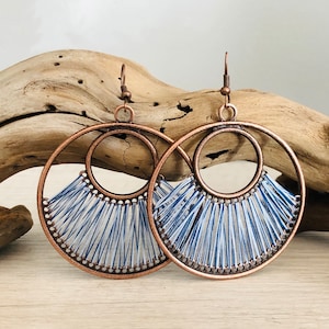 May include: A pair of large, copper-toned hoop earrings with a woven design in blue and white thread. The earrings have a circular shape with a smaller circle in the center.