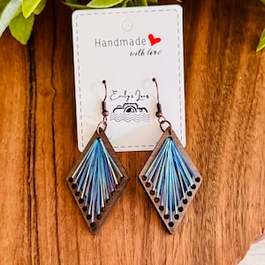 May include: Diamond-shaped wooden earrings featuring a dark brown frame with holes along the edges. Blue and iridescent thread is woven through the holes in a fan pattern. The earrings hang from copper-colored hooks on a white display card.