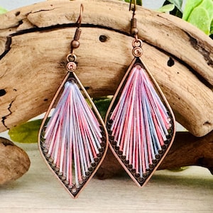 May include: A pair of copper-colored, diamond-shaped earrings. Each earring features a frame with a beaded edge and is filled with pink, white, and blue threads radiating from the center. The earrings hang from a simple hook.
