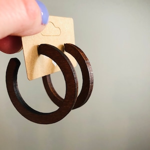 Puede incluir: Un par de pendientes de aro de madera de color marr&oacute;n oscuro sujetos a una tarjeta expositora rectangular de cart&oacute;n color canela. Los pendientes presentan un dise&ntilde;o circular de marco abierto con un acabado liso y pulido.