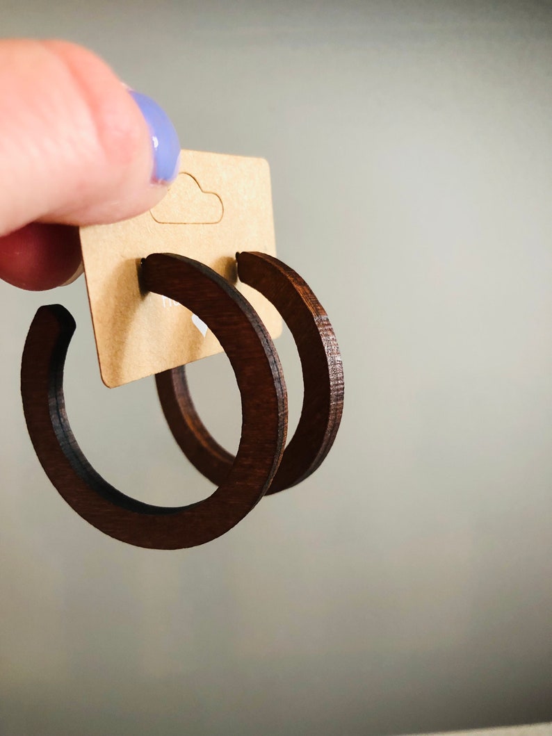 Puede incluir: Un par de pendientes de aro de madera de color marr&oacute;n oscuro sujetos a una tarjeta expositora rectangular de cart&oacute;n color canela. Los pendientes presentan un dise&ntilde;o circular de marco abierto con un acabado liso y pulido.