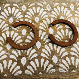 Puede incluir: Un par de pendientes de aro circulares hechos de madera de color marr&oacute;n oscuro. Cada pendiente presenta una superficie plana y lisa con un cierre de poste de metal plateado, dise&ntilde;ado para llevarse en el l&oacute;bulo con una estructura abierta.