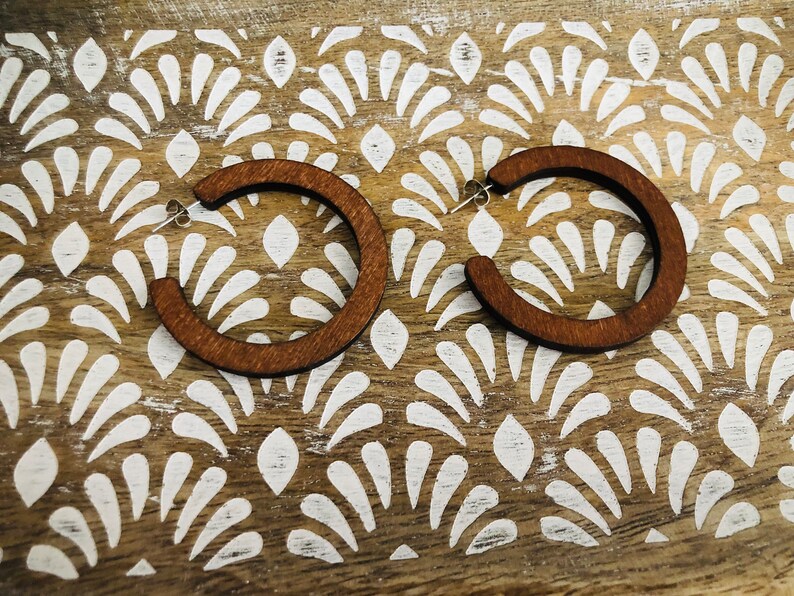 Puede incluir: Un par de pendientes de aro circulares hechos de madera de color marr&oacute;n oscuro. Cada pendiente presenta una superficie plana y lisa con un cierre de poste de metal plateado, dise&ntilde;ado para llevarse en el l&oacute;bulo con una estructura abierta.
