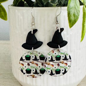 May include: A pair of Halloween-themed earrings. Each earring features a black witch hat and a circular charm with a repeating pattern of witch hats, cauldrons, brooms, and green potion bottles. The earrings hang from silver-colored hooks.