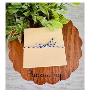Puede incluir: Una caja de regalo cuadrada de cart&oacute;n beige atada con cordel de panadero azul y blanco, centrada sobre una base de madera festoneada. La palabra Packaging est&aacute; impresa en negro en la parte inferior, con follaje verde arriba.