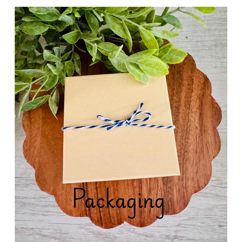 Puede incluir: Una caja de regalo cuadrada de cart&oacute;n beige atada con cordel de panadero azul y blanco, centrada sobre una base de madera festoneada. La palabra Packaging est&aacute; impresa en negro en la parte inferior, con follaje verde arriba.