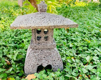 May include: A gray stone Japanese garden lantern with a square top and a rectangular base. The lantern has four circular openings on the front and is surrounded by green foliage. The lantern is approximately 46 cm tall.