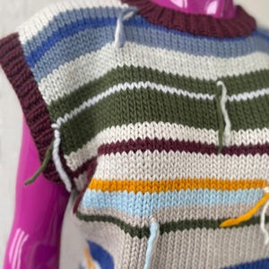 May include: A striped sleeveless sweater with a crew neck. The sweater is made of a soft, knitted fabric and features a variety of colors, including blue, green, white, brown, yellow, and orange. The sweater has a unique design with long, dangling threads at the edges.