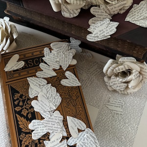 May include: A collection of white paper roses made from book pages. The roses are arranged on a stack of old books and a vintage book with a gold-embossed design. The roses are made from pages with text, creating a unique and artistic look.