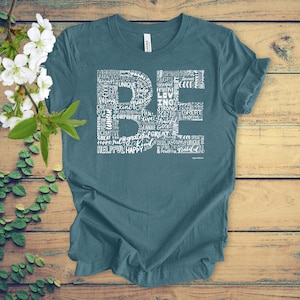 Puede incluir: Camiseta azul turquesa con un dise&ntilde;o gr&aacute;fico blanco que deletrea "BE" usando palabras positivas como "feliz", "amor", "fuerte", "amable" y "agradecido".
