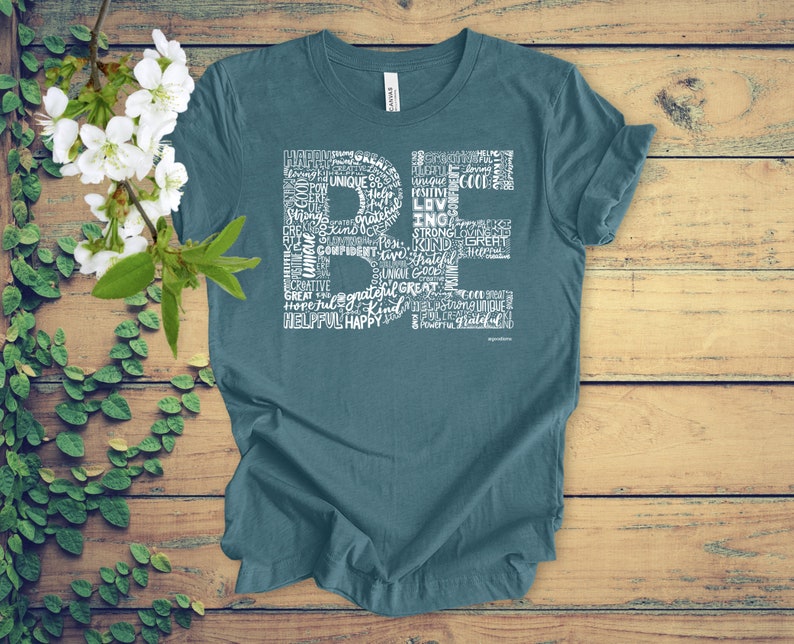 Puede incluir: Camiseta azul turquesa con un dise&ntilde;o gr&aacute;fico blanco que deletrea "BE" usando palabras positivas como "feliz", "amor", "fuerte", "amable" y "agradecido".
